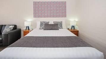A neatly made bed with a striped white and gray duvet, two bedside lamps, a black leather sofa, and a floral-patterned wall hanging.