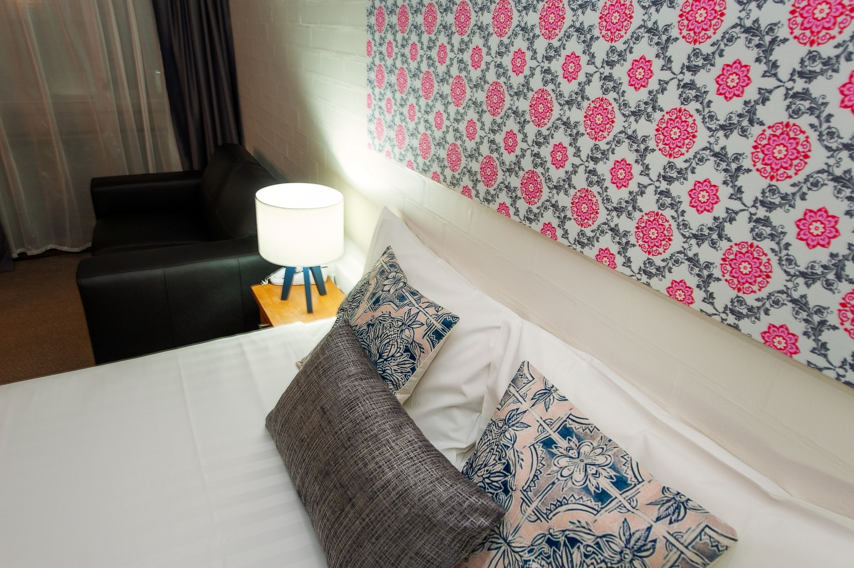 Hotellihuone, jossa on sänky, tyynyjä, yöpöytä, lamppu ja kuviollinen tapetti.