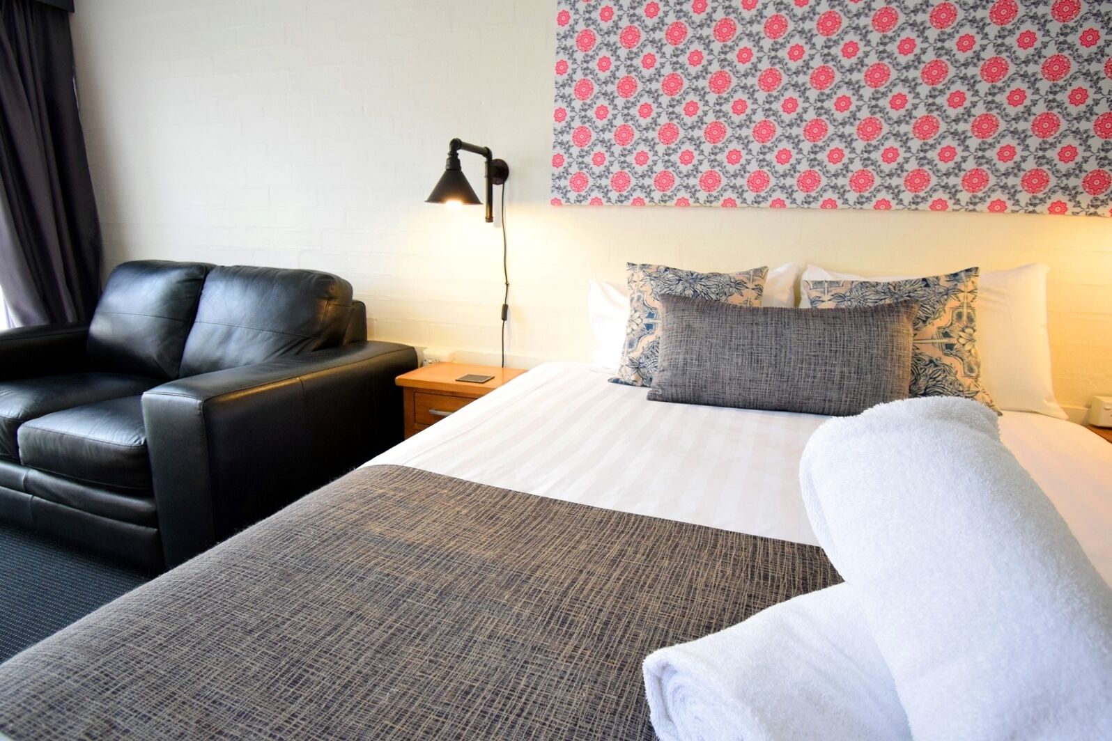 A hotel room with a bed, a chair, a bedside table, a lamp, and patterned wallpaper.