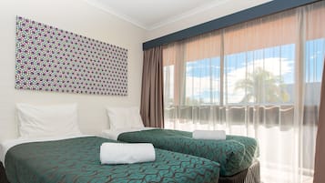 A hotel room with two beds, a patterned wall hanging, and a large window with sheer curtains.