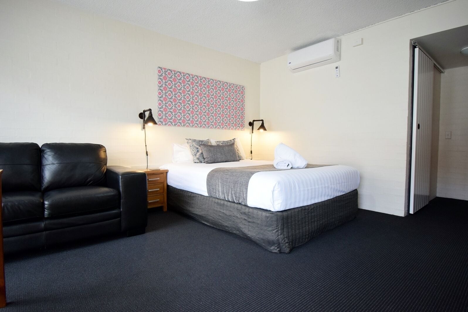 A hotel room with a bed, a black leather chair, a nightstand, two bedside lamps, and a patterned wall hanging.