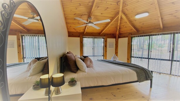 A spacious bedroom with a large bed, two bedside tables with lamps, a ceiling fan, and large windows with blinds.