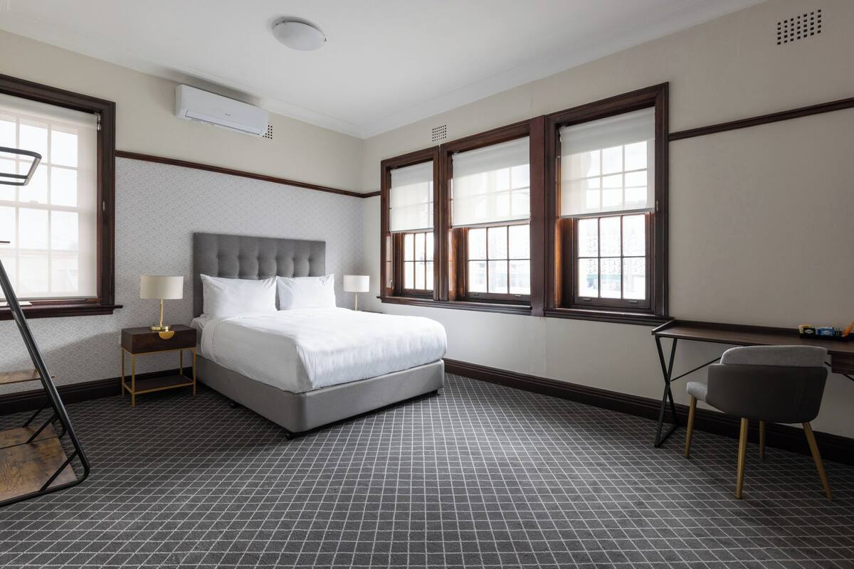 A hotel room with a bed, two bedside tables, a desk, a chair, and large windows.