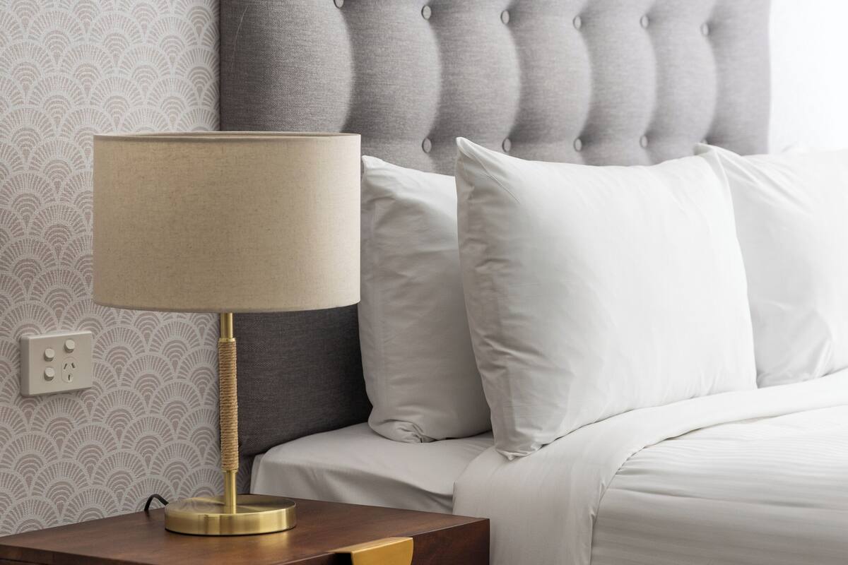 A neatly made bed with a tufted headboard, a bedside table with a lamp, and a patterned wallpaper.