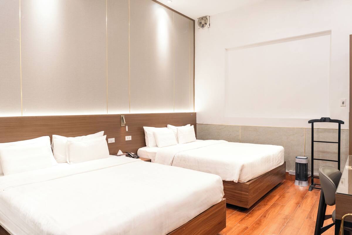 A hotel room with two beds, a desk, a chair, a heater, and a wall-mounted fan.