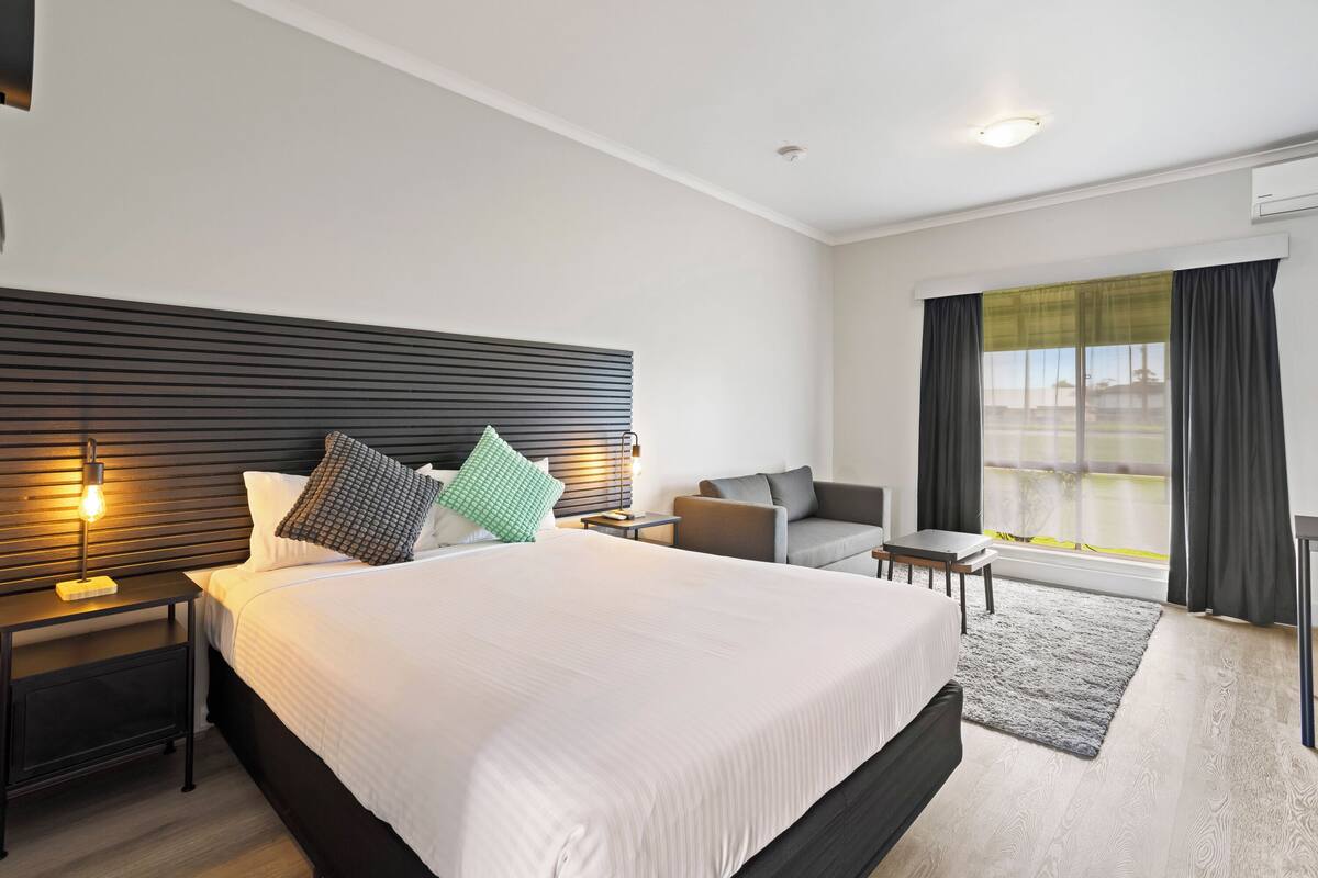 Une chambre d’hôtel moderne, dotée d’un grand lit, d’une table de chevet, d’un bureau, d’une chaise et offrant une vue sur l’extérieur.