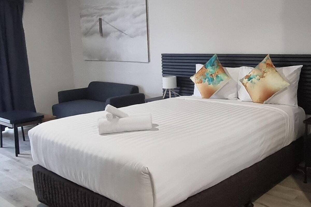 Un lit bien fait, avec une couette blanche et des oreillers, une tête de lit sombre, une table de chevet et une horloge fixée au mur.