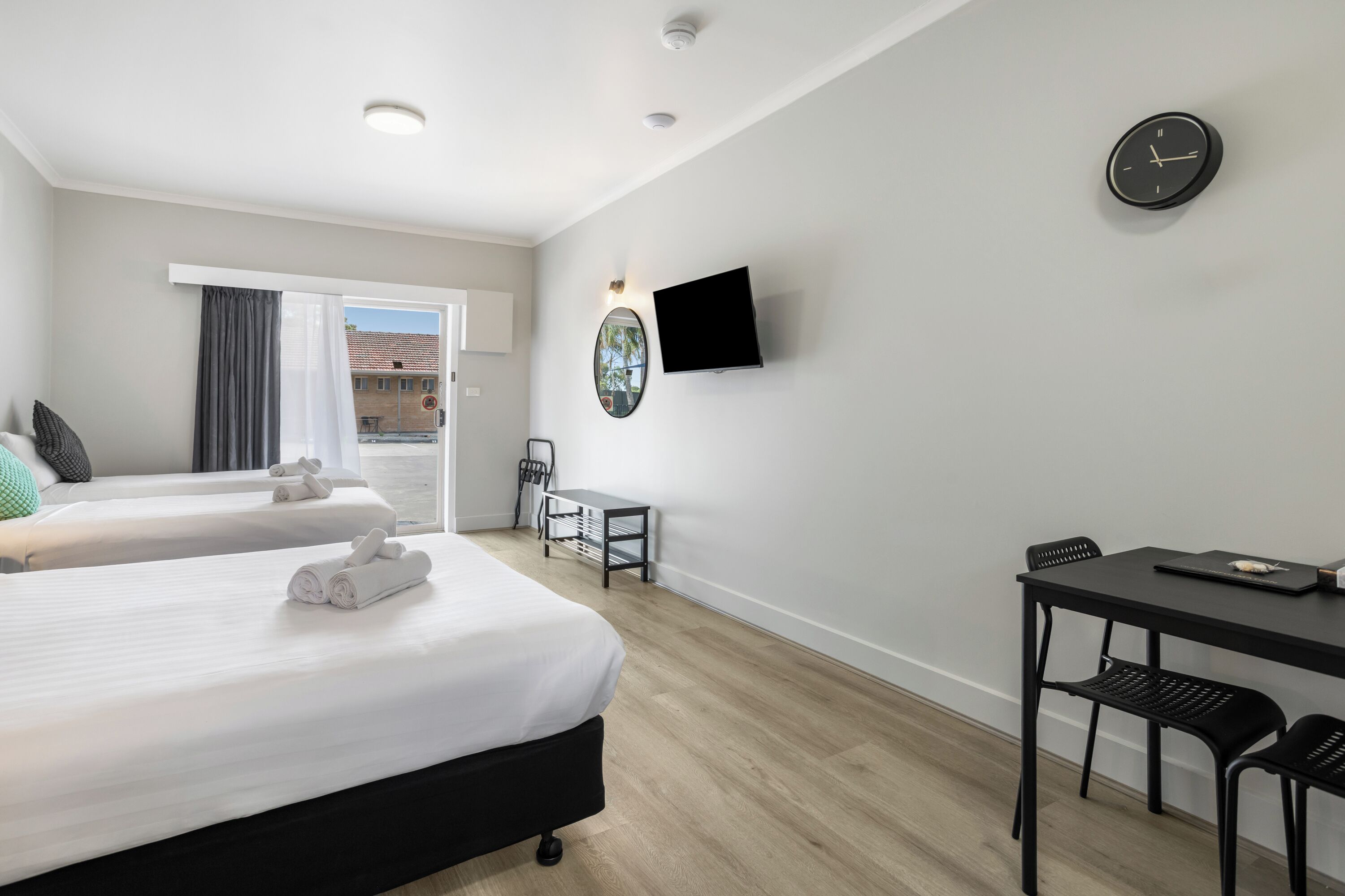 A hotel room with two beds, a desk, a chair, a TV, and a clock.