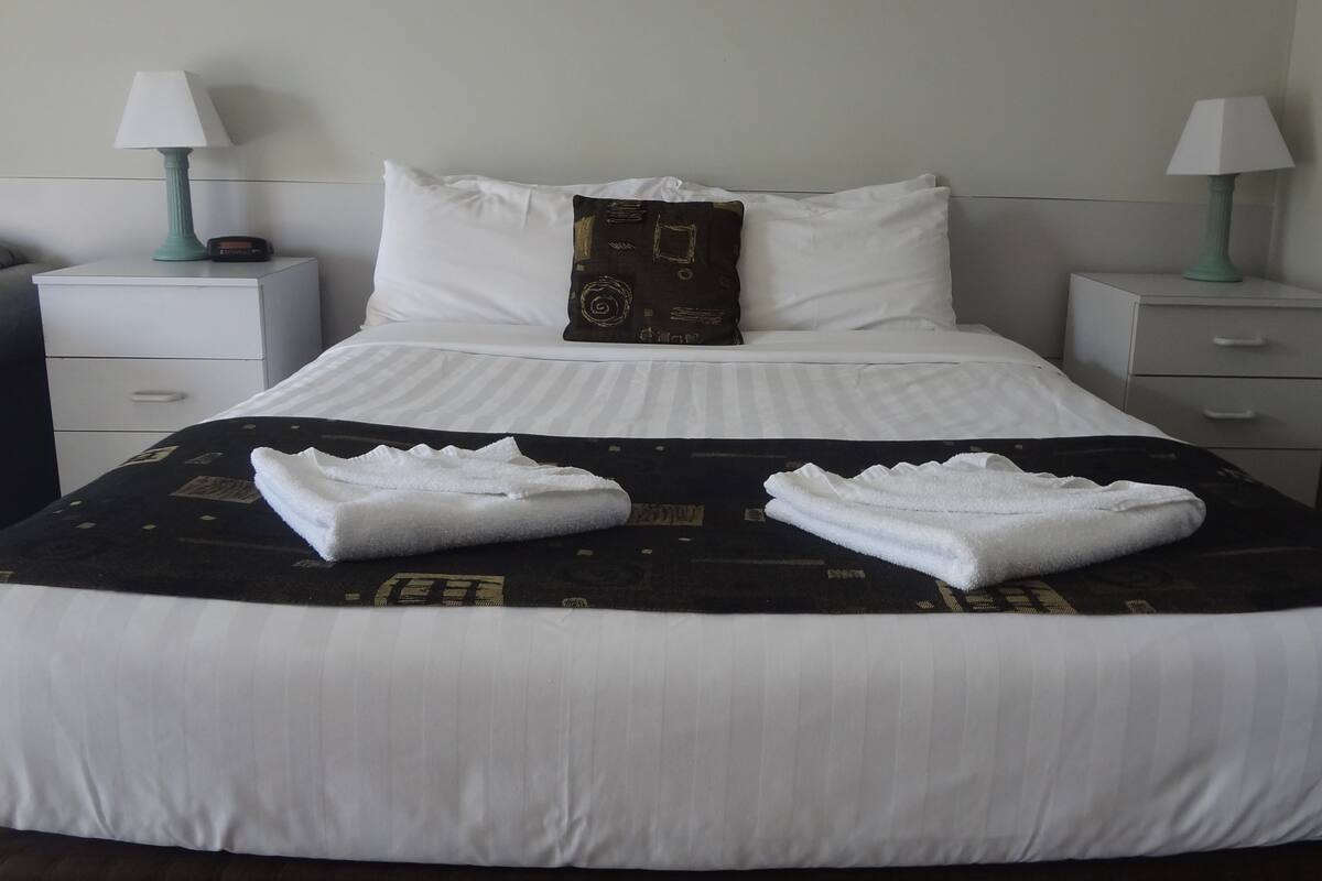 Un lit bien fait, avec du linge de lit blanc, un plaid noir à motifs et deux serviettes pliées. On y trouve deux tables de chevet avec des lampes, un fauteuil gris et une commode blanche.