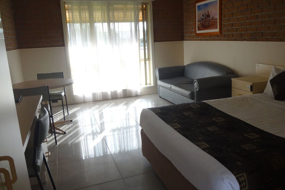 Habitación de hotel con cama, escritorio, silla, sofá, mesita de noche y ventana con cortinas.