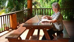 A person using a laptop at a wooden picnic table outdoors with a railing and greenery in the background. - Atherton Holiday Park (Atherton)