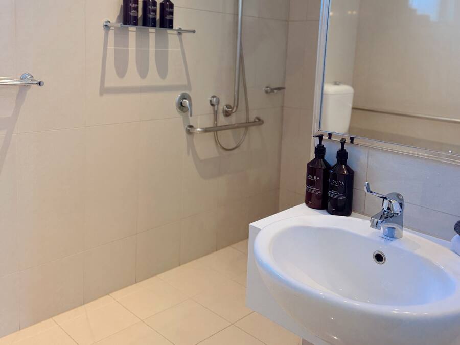 Un baño con lavamanos blanco, un espejo, ducha y artículos de aseo en un estante.
