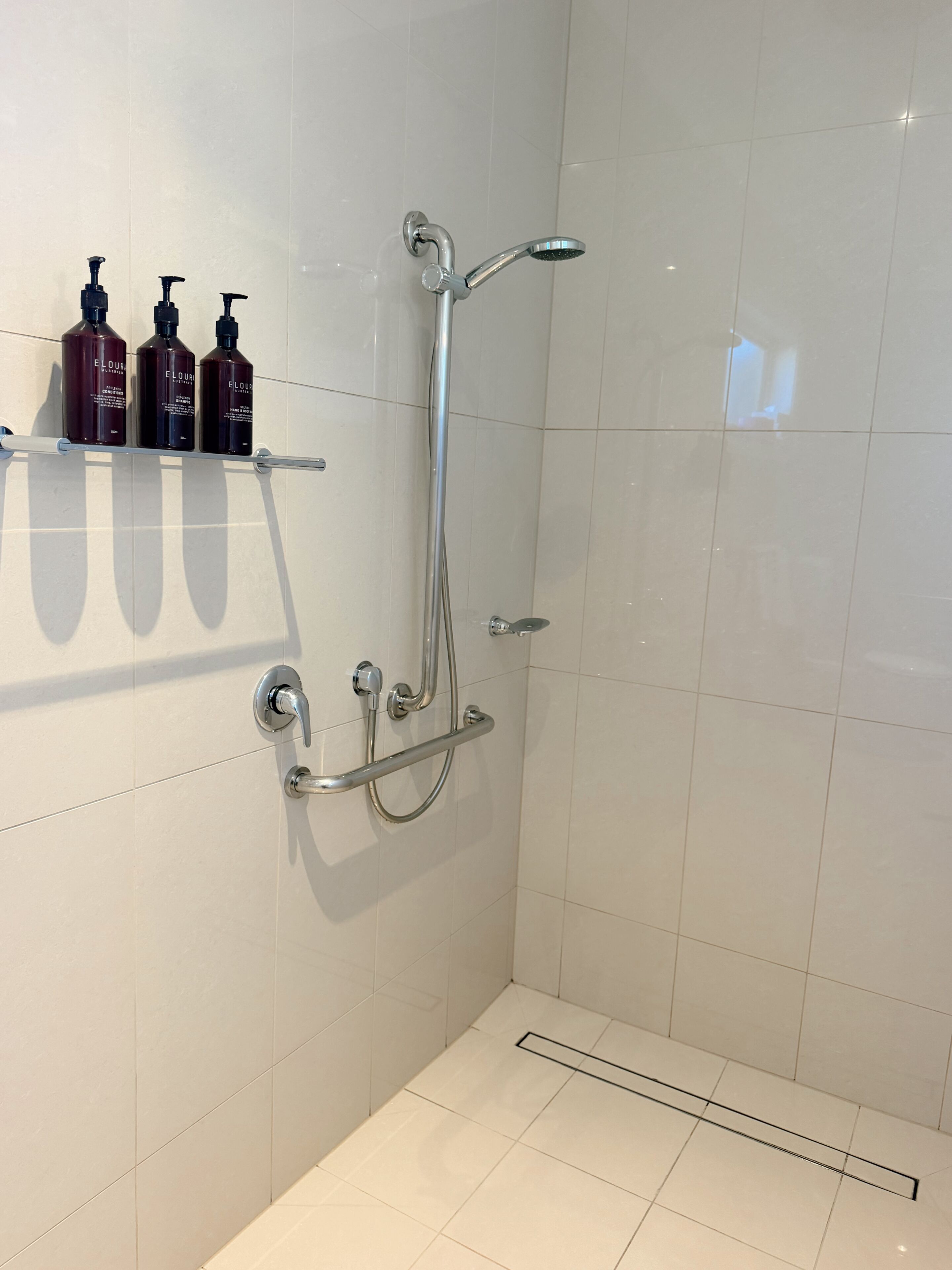 Eine Dusche mit Handbrause und Handtucharmatur, eine Duschkabinentür aus Glas und ein Ablagefach mit drei Flaschen Elour Körperlotion.