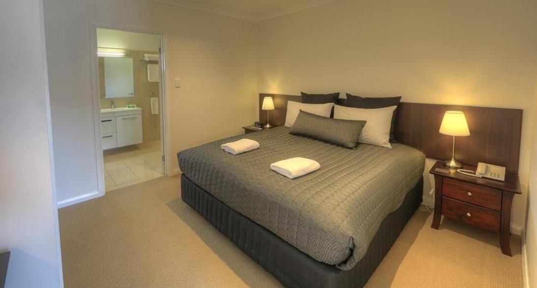 A neatly arranged hotel room with a large bed, two bedside tables with lamps, a small bedside table with a phone, and a bathroom visible through an open door.