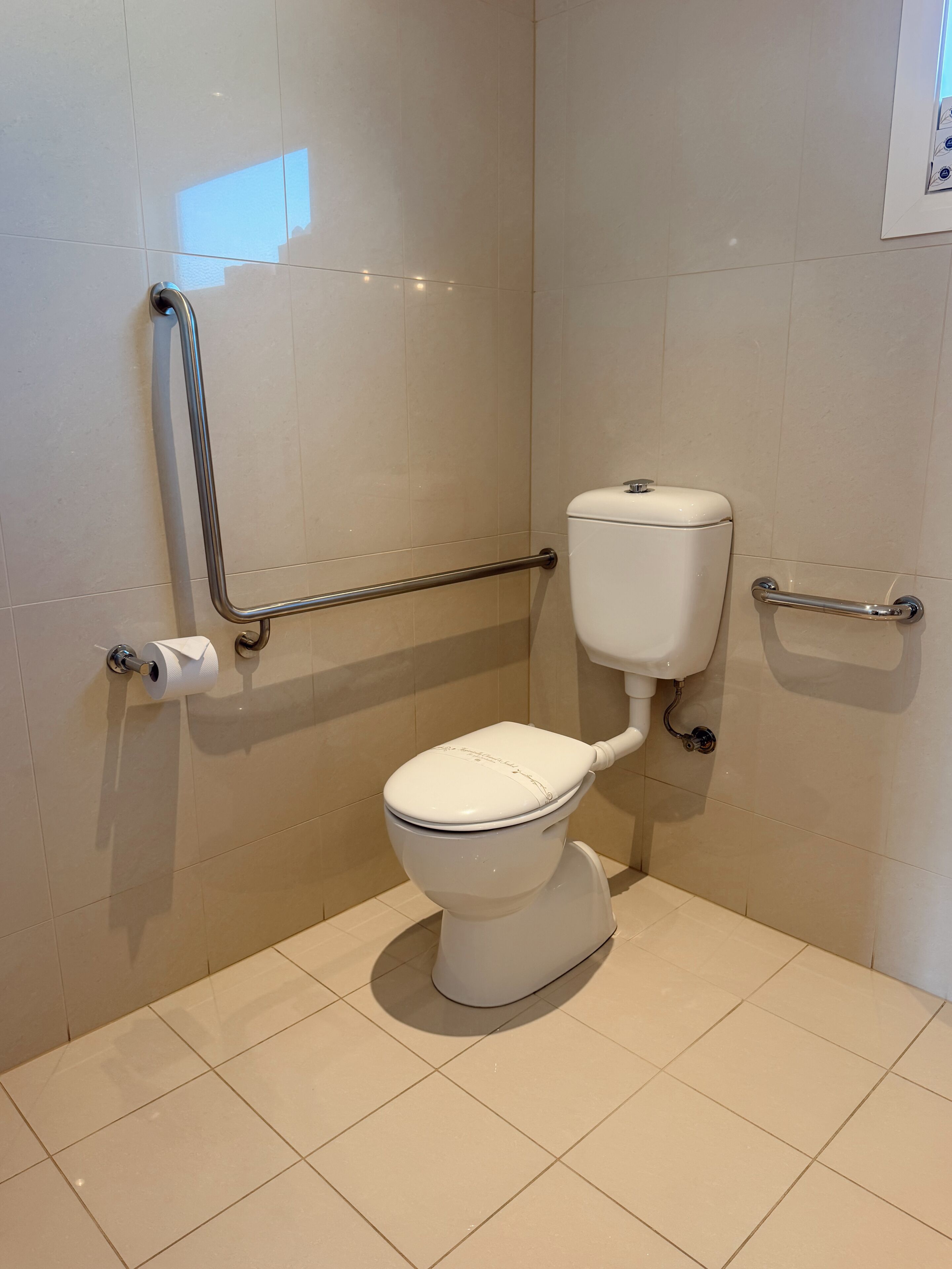 Sanitário branco com barras de apoio, rolo de papel higiénico e chão em azulejos.