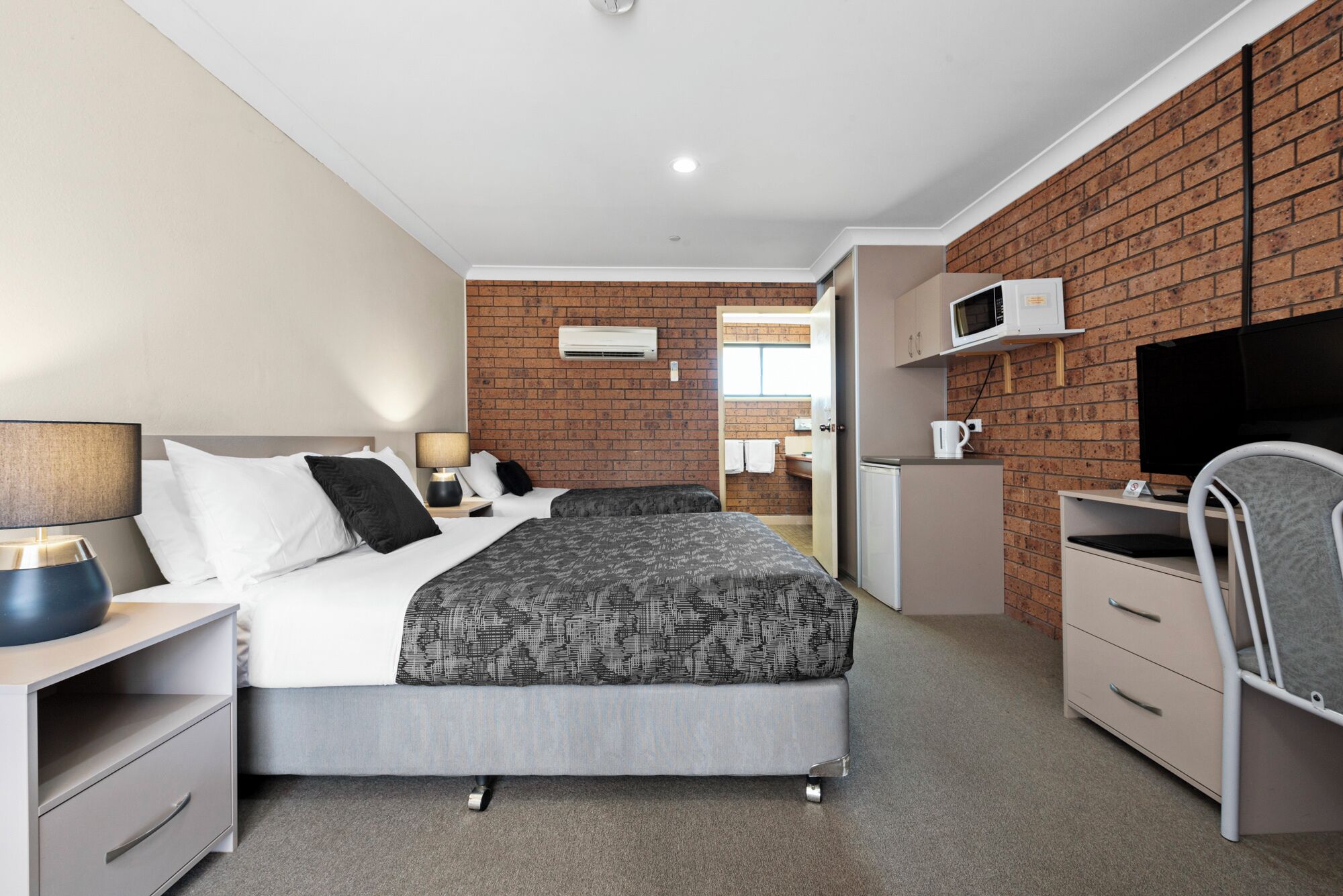 A modern hotel room with a brick wall, a bed with grey bedding, a desk with a chair, a microwave, and a TV.
