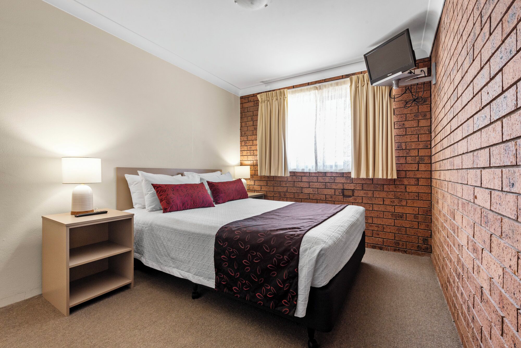 A hotel room with a brick wall, a bed with white bedding and red pillows, a bedside table with a lamp, and a TV mounted on the wall.