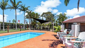 Una piscina con plataforma de ladrillo, rodeada de palmeras y una zona sombreada con sillas de descanso blancas.