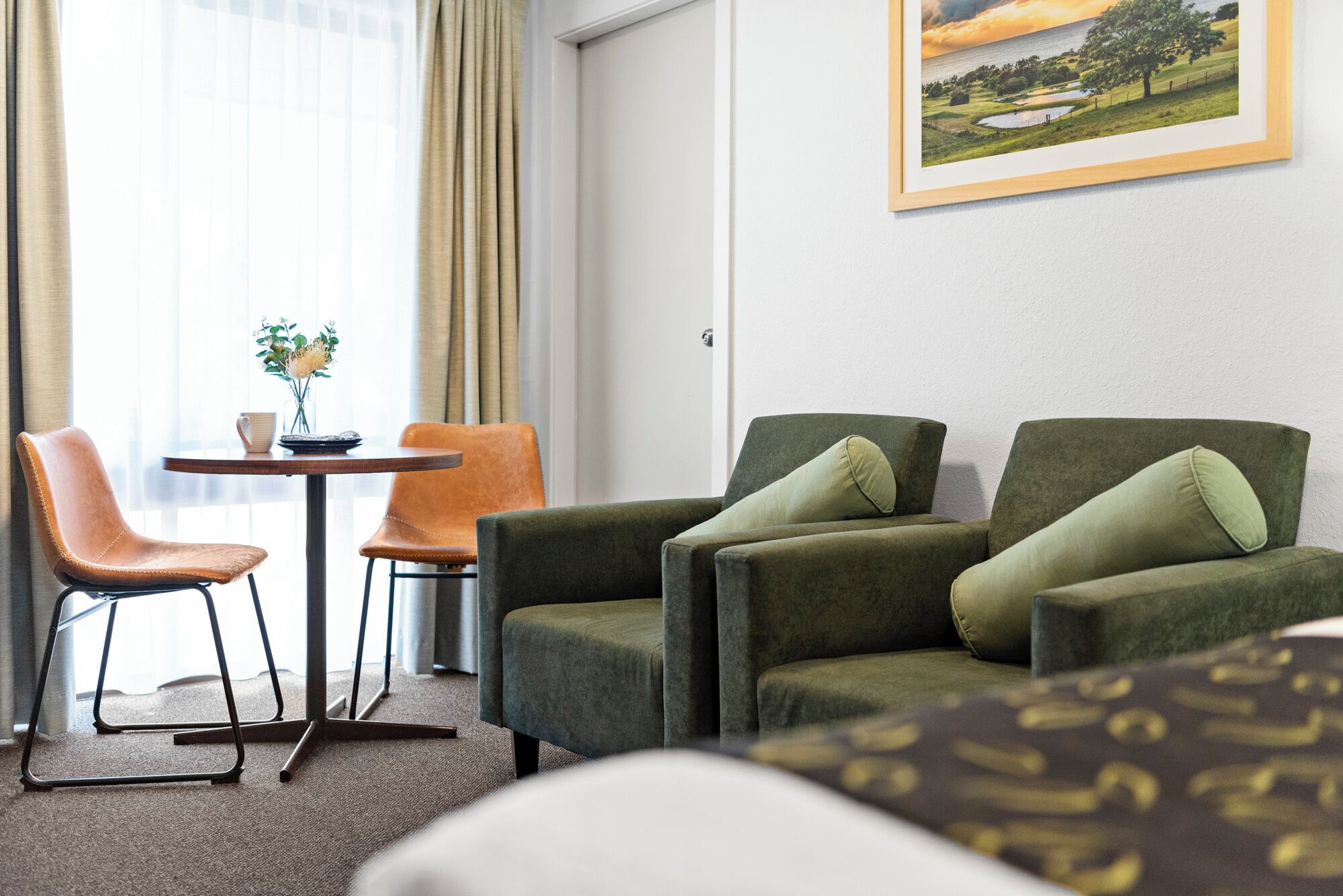 Habitación de hotel con sofá verde, una butaca de cuero marrón, una mesita redonda con un florero y un cuadro de paisaje en la pared.