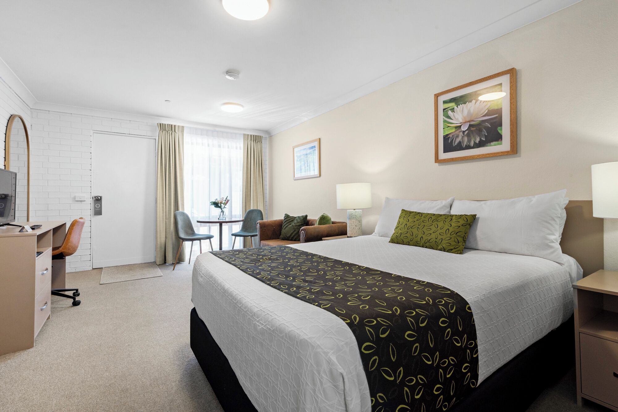 A hotel room with a bed, a desk with a chair, a small table with two chairs, a lamp, and a framed picture on the wall.