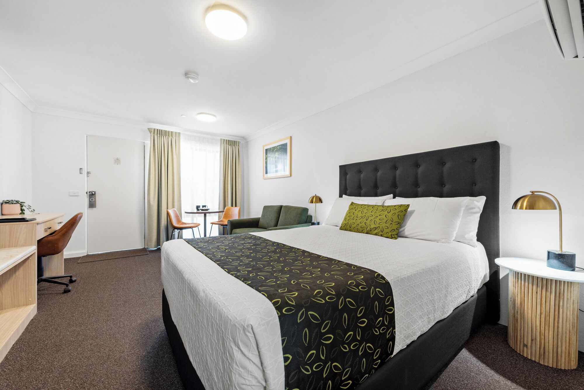 A hotel room with a large bed, a desk with a chair, a small table, and a lamp.