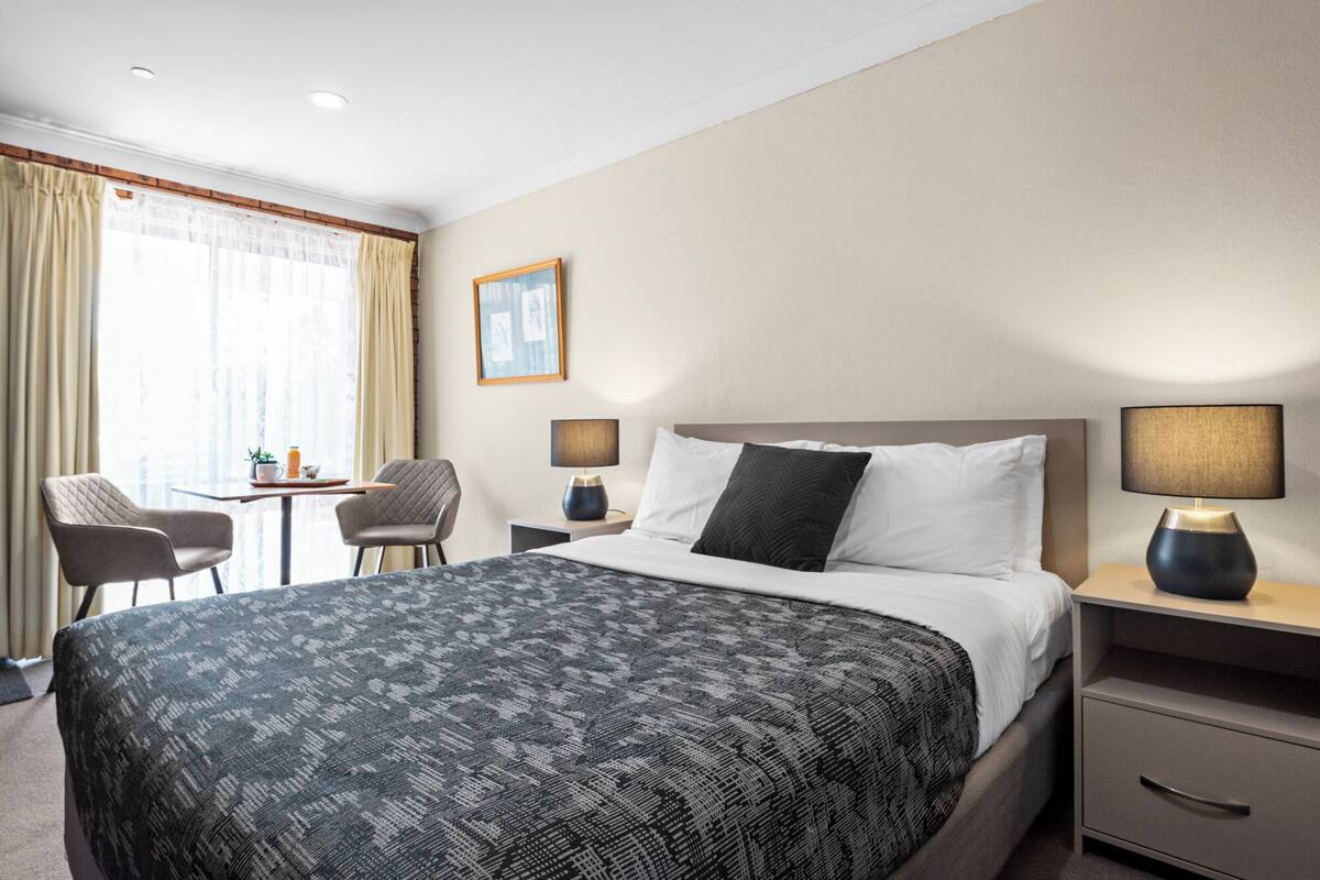A hotel room with a bed, bedside tables, a chair, a small table with a plant, and a window with curtains.