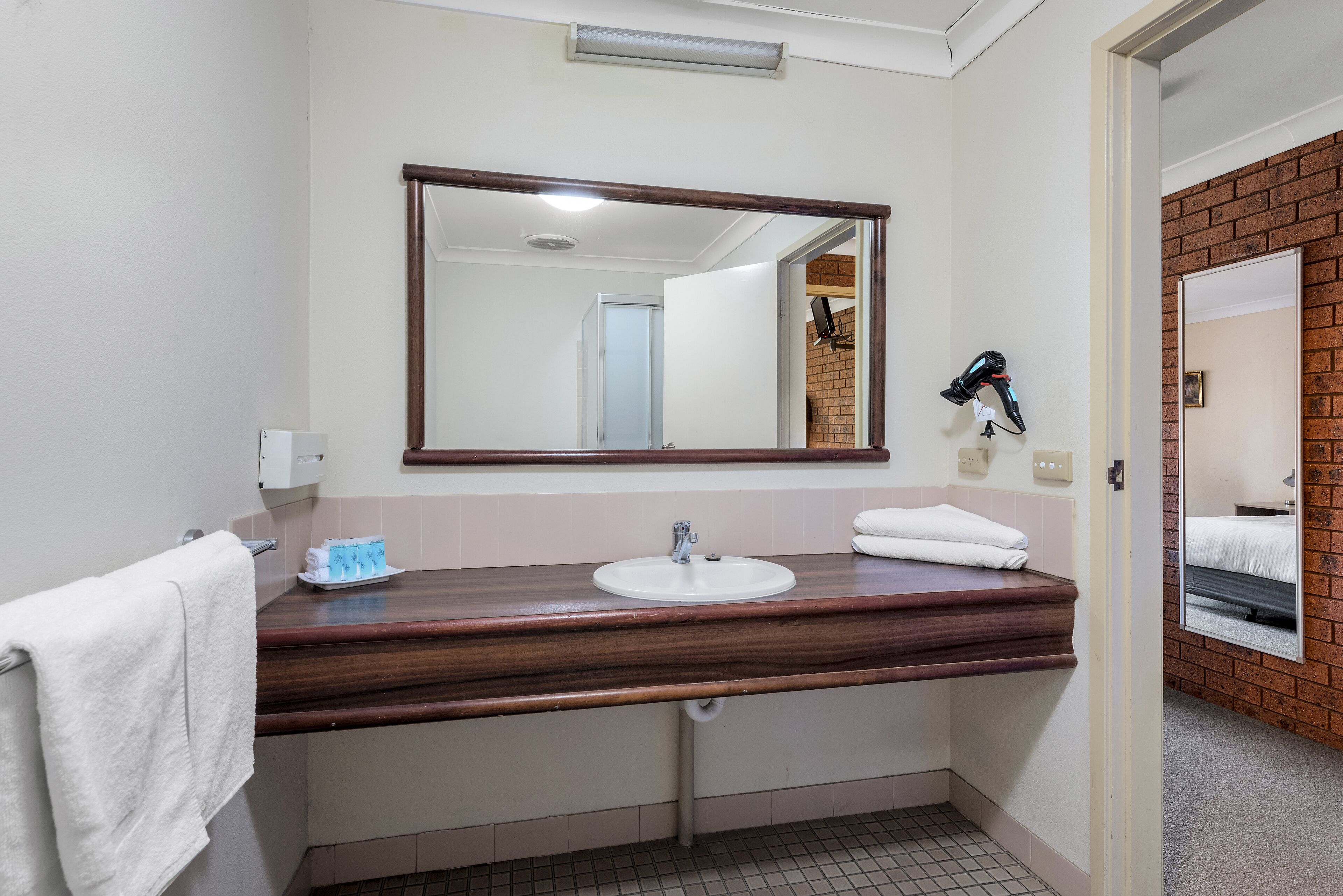 A bathroom with a wooden vanity, a white sink, a mirror, and a hairdryer on the wall.
