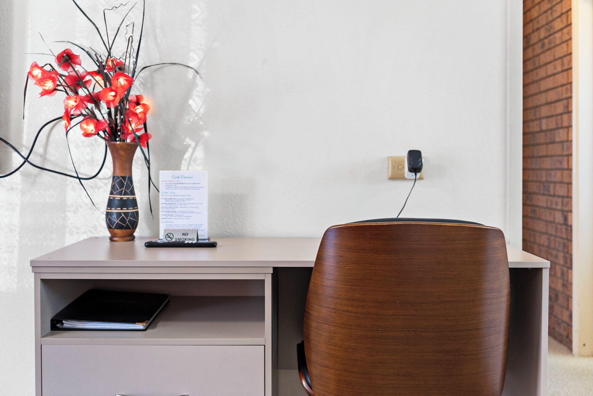 Un bureau avec un ordinateur portable, un vase de fleurs rouges, un panneau «interdiction de fumer» et une prise électrique sur un mur.