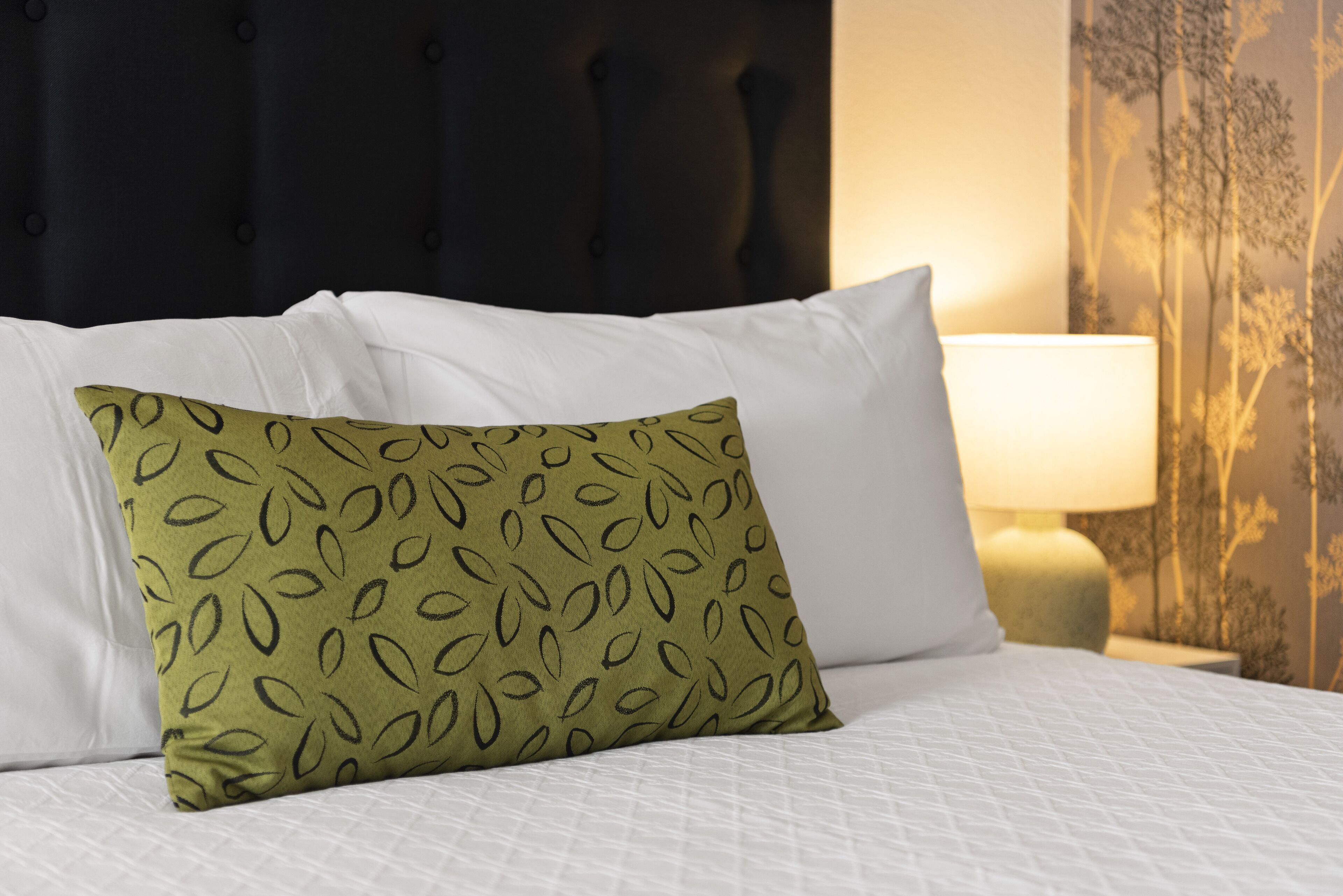 Una cama con ropa de cama blanca, un cojín estampado en verde, un cabecero oscuro y una lámpara en una mesita de noche.