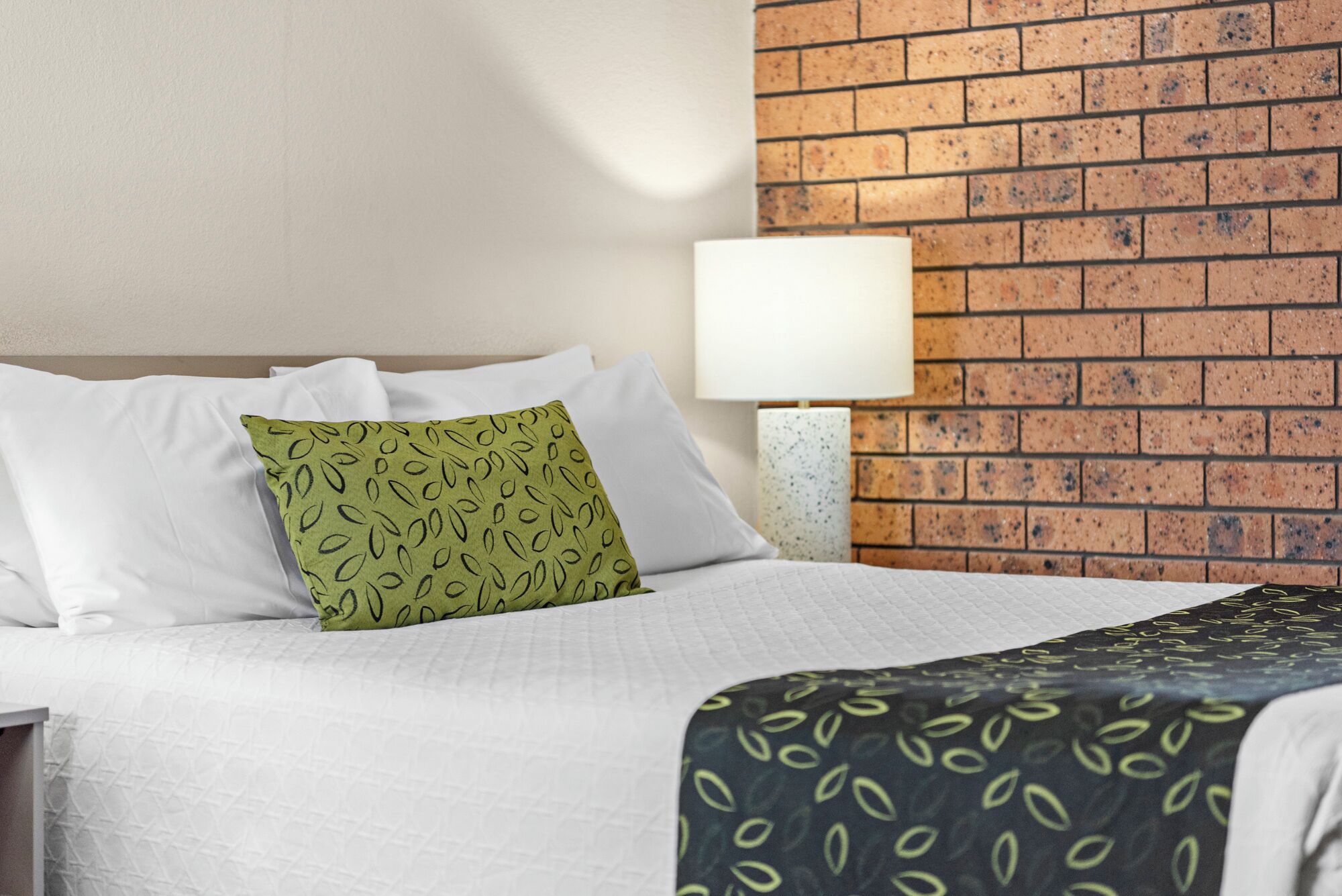 A hotel room with a brick wall, a bed with white bedding, a green pillow with a leaf pattern, and a black and green patterned throw.