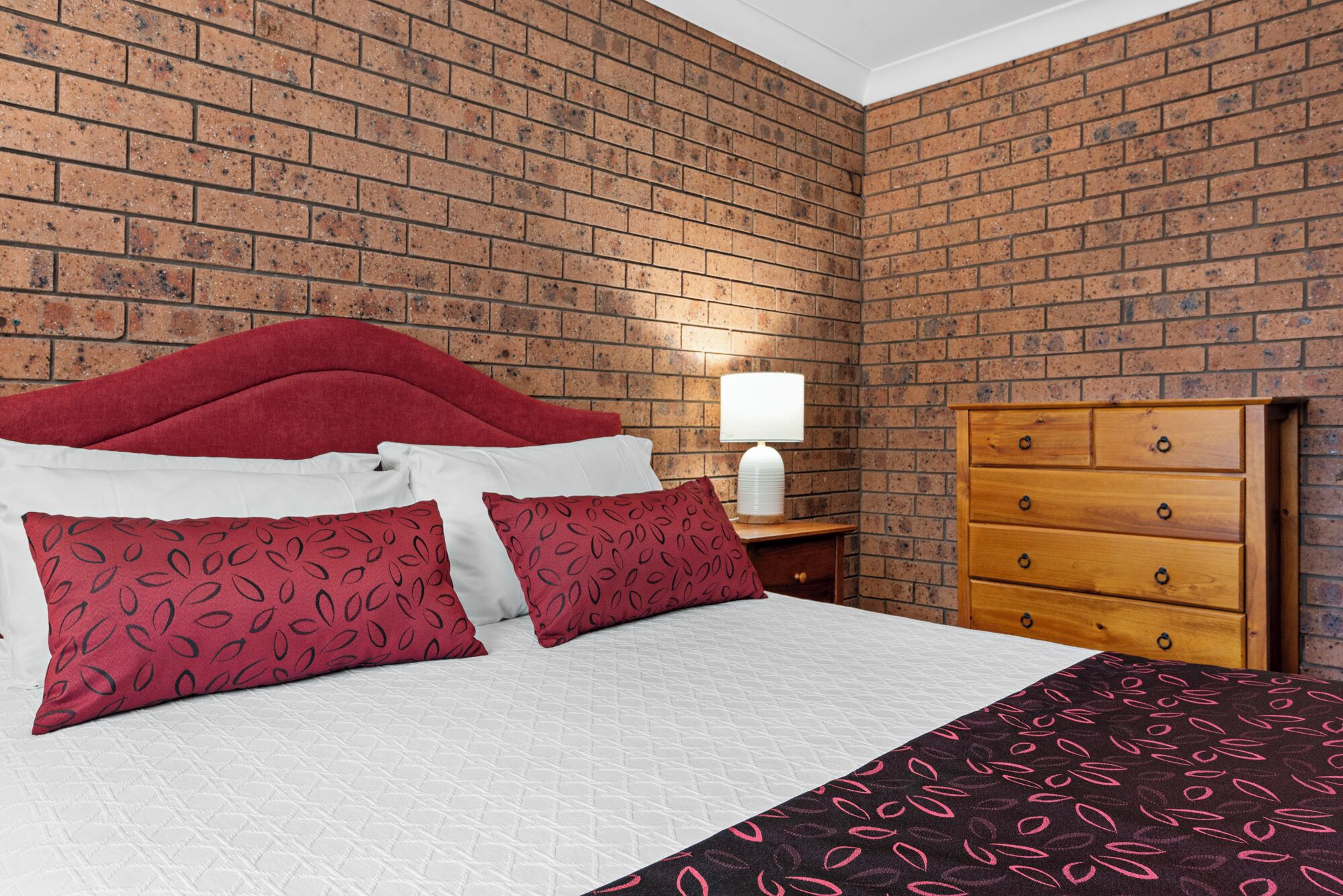 A bedroom with a brick-patterned wall, a bed with red and white bedding, a wooden dresser, and a bedside lamp.