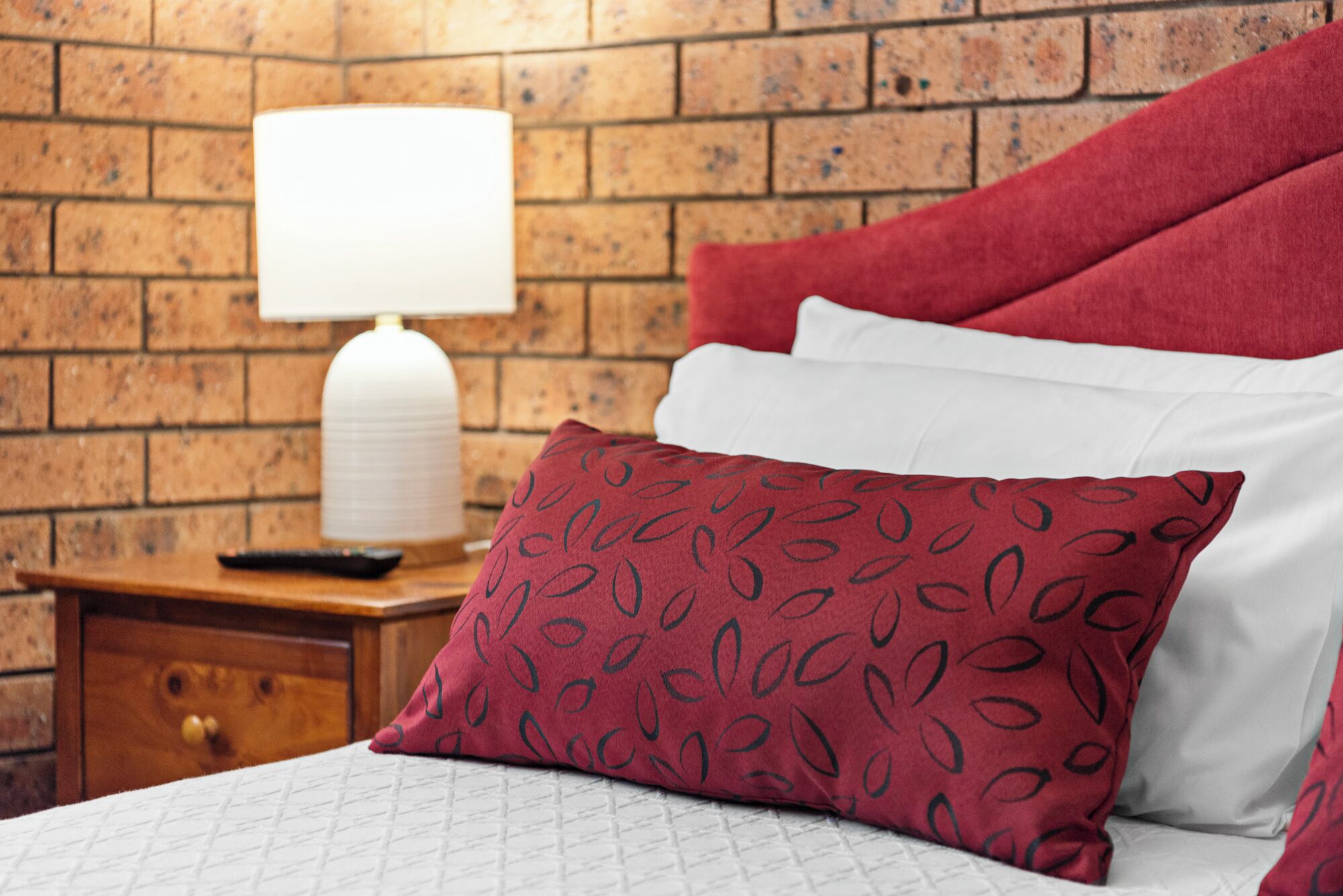 Un lit bien fait, agrémenté d’un coussin à motifs, d’un coussin blanc et d’un plaid rouge. Une table de chevet en bois, sur laquelle se trouvent une lampe et une télécommande, est placée à côté du lit.