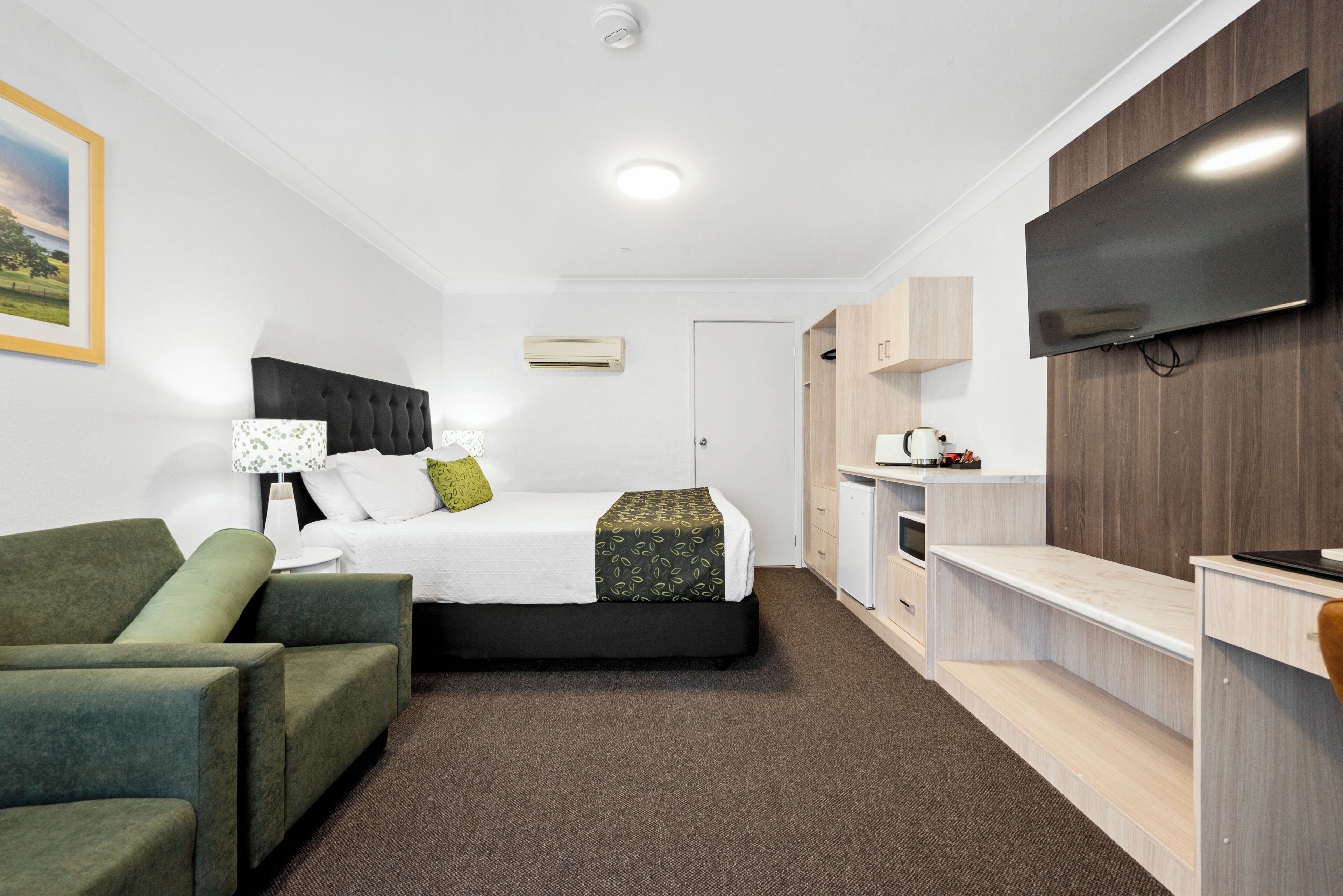 Una habitación de hotel moderna con cama, sillón verde, televisor y una zona de kitchenette.