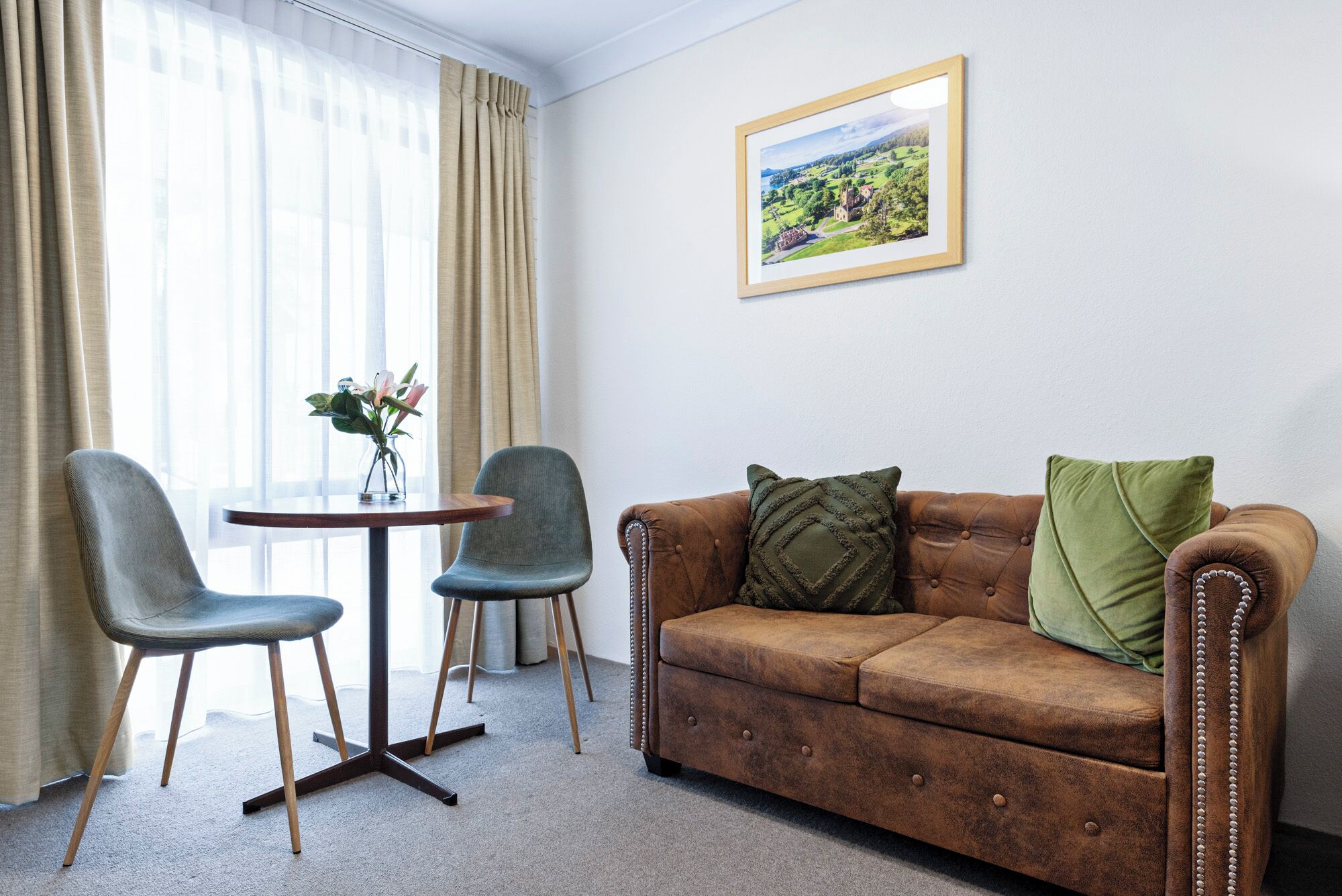 A room with a brown leather sofa, two grey chairs, a small round table, and a framed landscape painting on the wall.