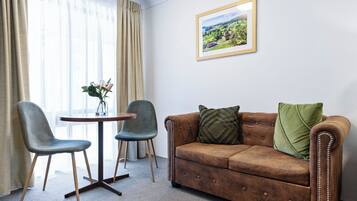 A room with a brown leather sofa, two grey chairs, a small round table, and a framed landscape painting on the wall.