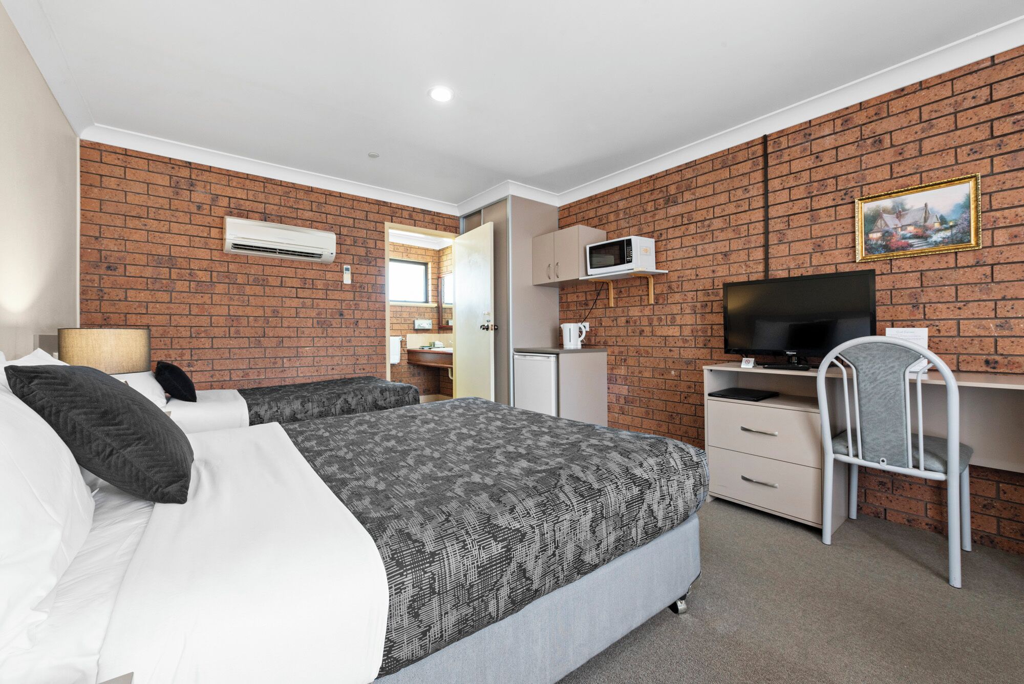 A hotel room with a brick wall, a bed with patterned bedding, a desk with a chair, a television, and a microwave.