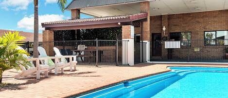 Una piscina con un porche cubierto, sillas de descanso y un edificio de ladrillo con techo metálico.