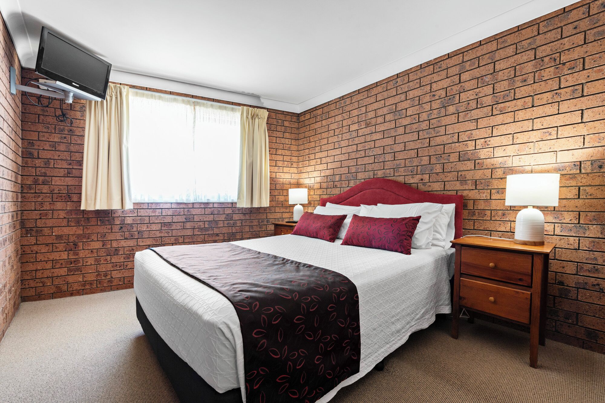 Een hotelkamer met een muur in baksteenmotief, een bed met een rode hoofdbord, twee nachtlampjes en een aan de muur bevestigde televisie.