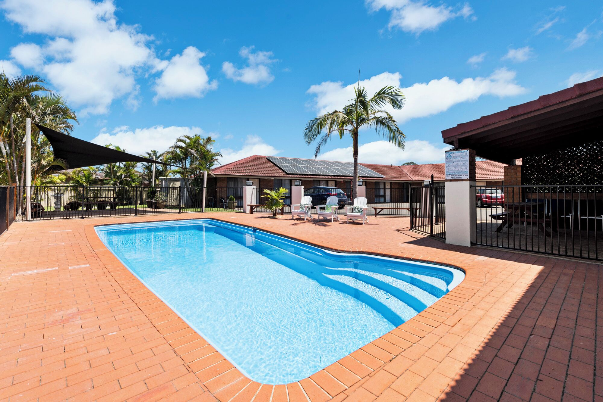 Una piscina rectangular con borde de ladrillo, rodeada por un patio cercado con sillones de descanso y una zona de estar cubierta.