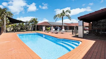 Una piscina rectangular con borde de ladrillo, rodeada por un patio cercado con sillones de descanso y una zona de estar cubierta.