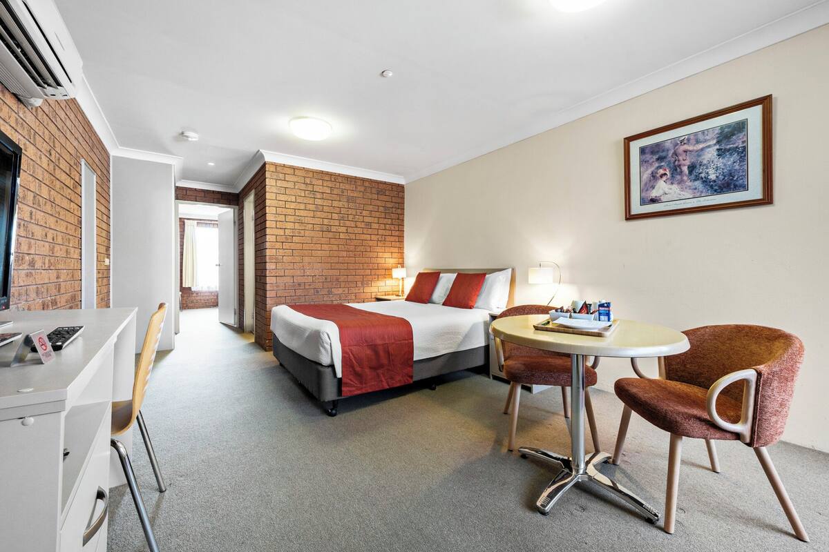 A hotel room with a bed, a desk, a chair, a table, and a painting on the wall.
