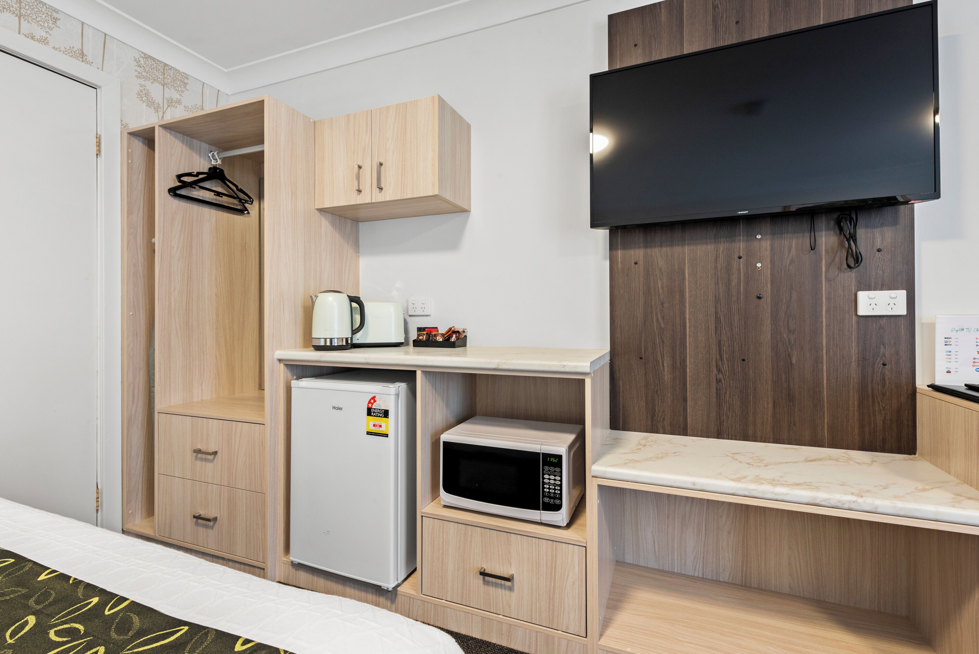 Habitación de hotel con cama, armario, refrigerador pequeño, microondas, cafetera y televisor de pantalla plana montado en la pared.