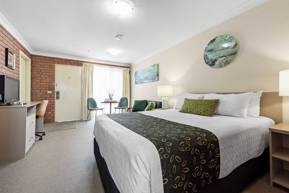 A hotel room with a bed, desk, chair, and a small table with a lamp.