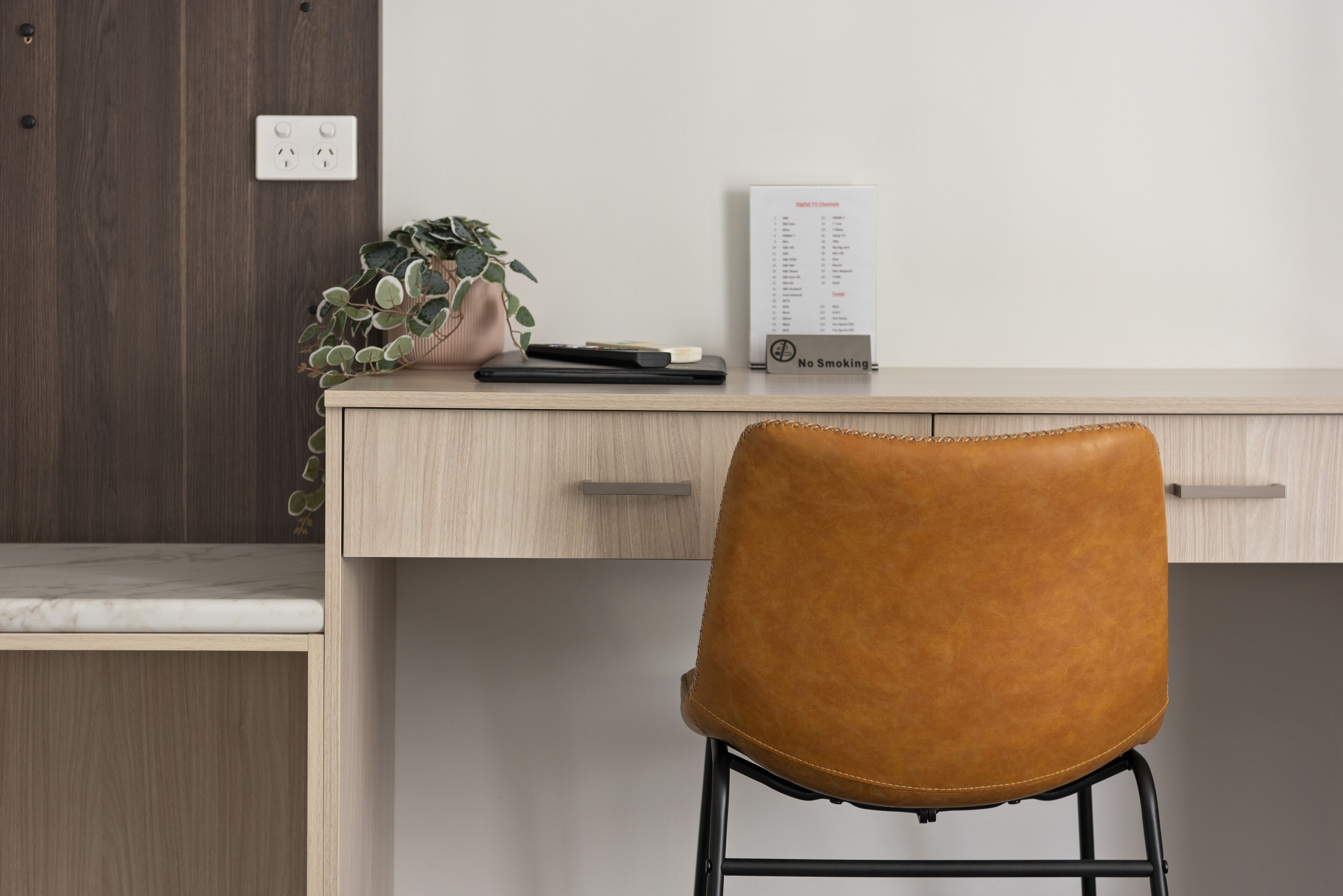 Un espacio de trabajo con un escritorio, una silla de cuero marrón, una planta en maceta, una libreta negra y un cartel de 'No Fumar'.