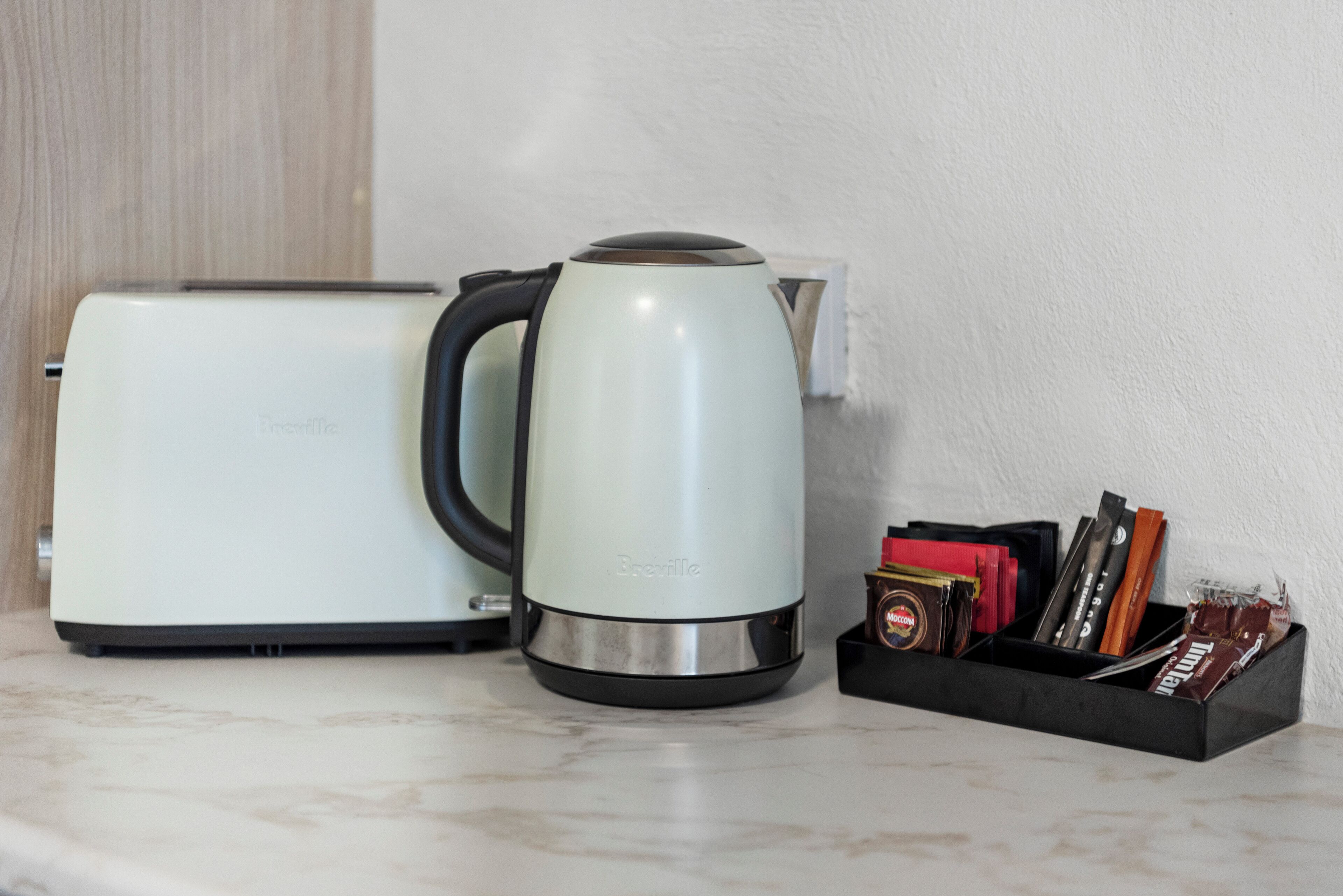 Un grille-pain et une bouilloire de la marque Breville sont posés sur un plan de travail, accompagnés d’un plateau noir contenant divers objets.