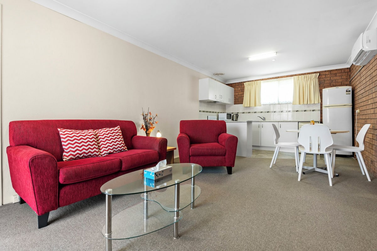 A compact living space with a red sofa, a glass coffee table, a red armchair, and a small dining area with white chairs. A kitchenette with a microwave and a refrigerator is visible in the background.