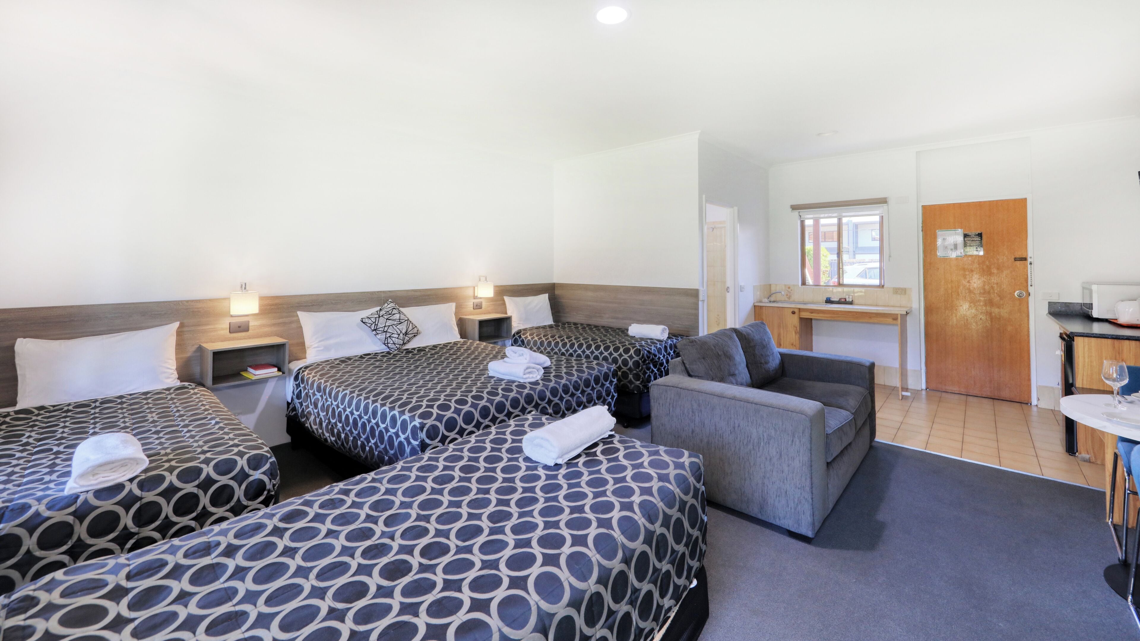 A hotel room with three beds, a sofa, a small kitchenette, and a door leading to the outside.