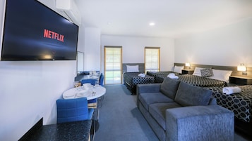 A hotel room with a TV displaying the Netflix logo, a sofa, two beds, a dining area with a table set for two, and a window.