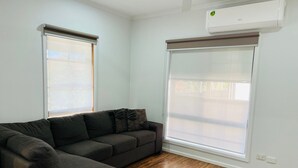 A modern living room with a sectional sofa, large windows with blinds, a ceiling air conditioner, and hardwood flooring. - The Cottage Motor Inn Albury CBD (South Albury)