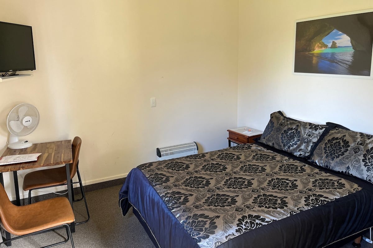 Ein Hotelzimmer mit einem Bett, einem Schreibtisch mit Stuhl, einem Standventilator, einem Heizgerät und einem Gemälde einer Küstenlandschaft.